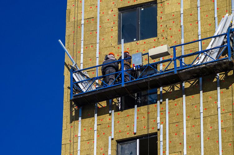 Two workmen in a gondola on the side of a tower block installing cladding.