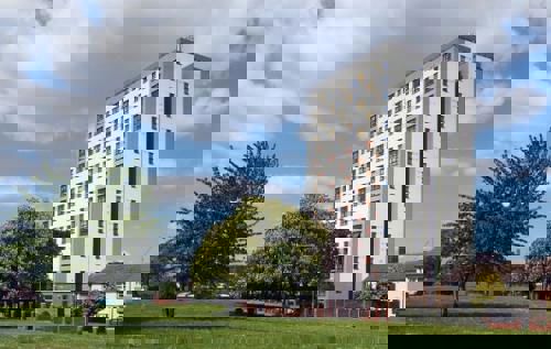 A view of an 18 storey tower block next to a house with a park of grass and trees in the foreground.