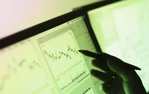 A pair of monitor screens showing complex dashboards of data as graphs. A hand holds a pen and is pointing at one graph.